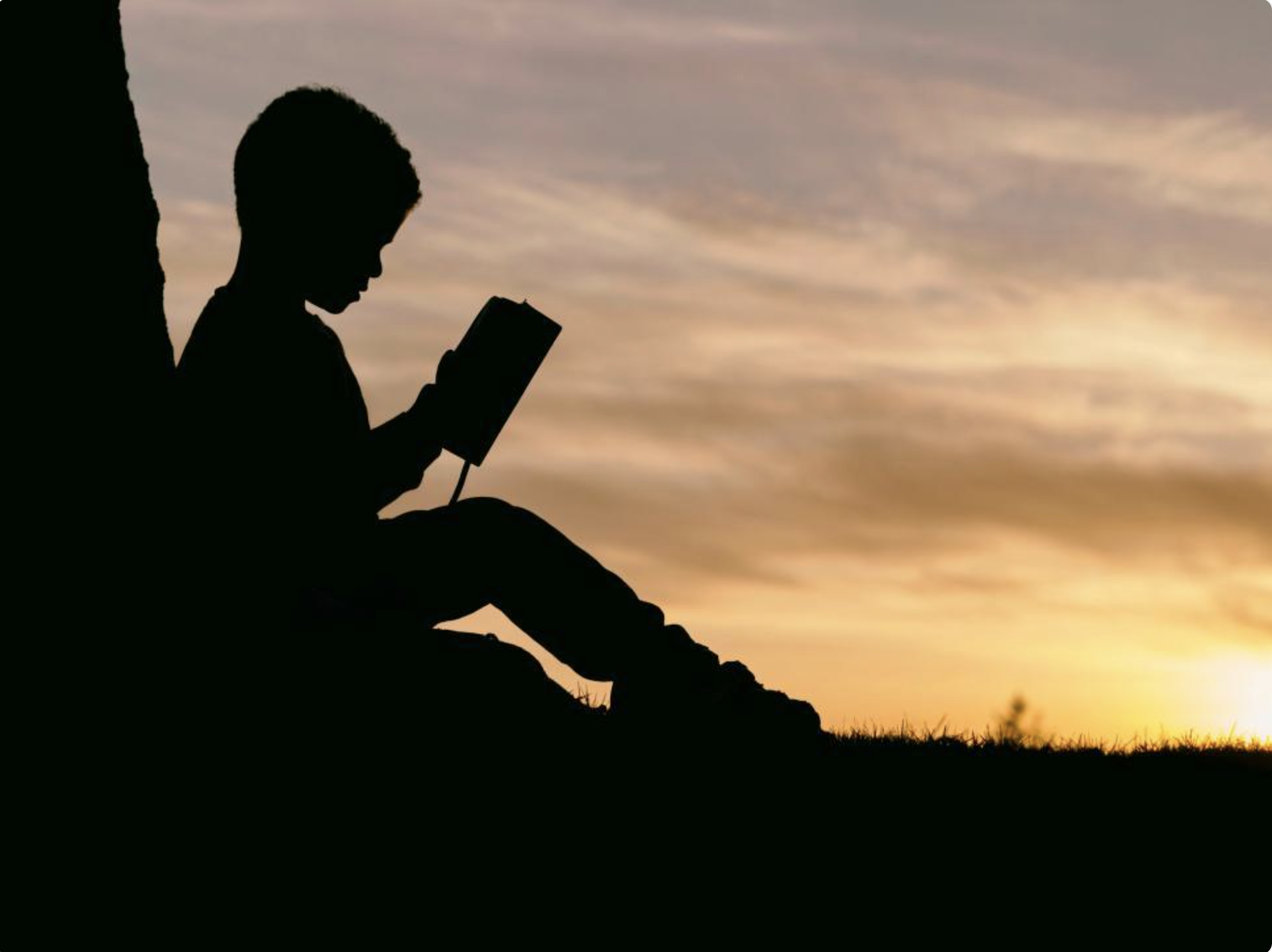 Silhouette of a child reading a book against a sunset sky